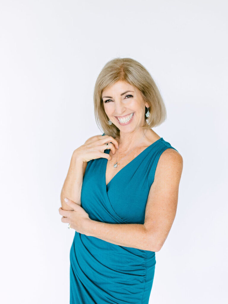 Lora Ulrich, laughing lightly, poses in a teal dress, touching her necklace