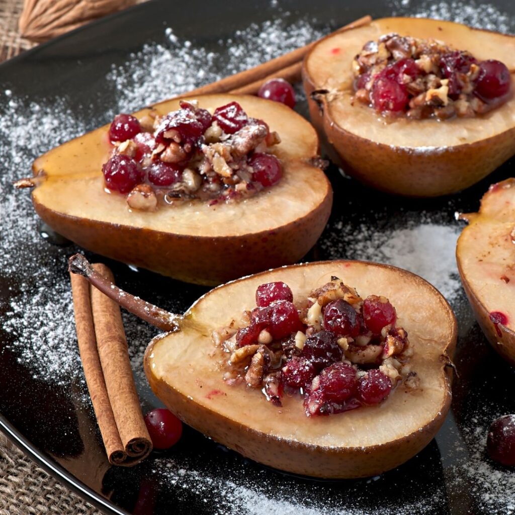 Baked pear halves filled with chopped walnuts and cranberries, garnished with cinnamon sticks on a dark plate.