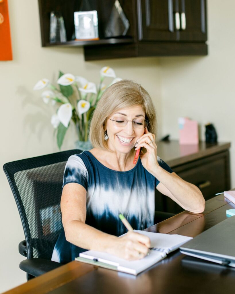 A certified holistic nutritionist speaking with a midlife client over the phone, taking notes during a free clarity call, offering personalized, educational wellness support.