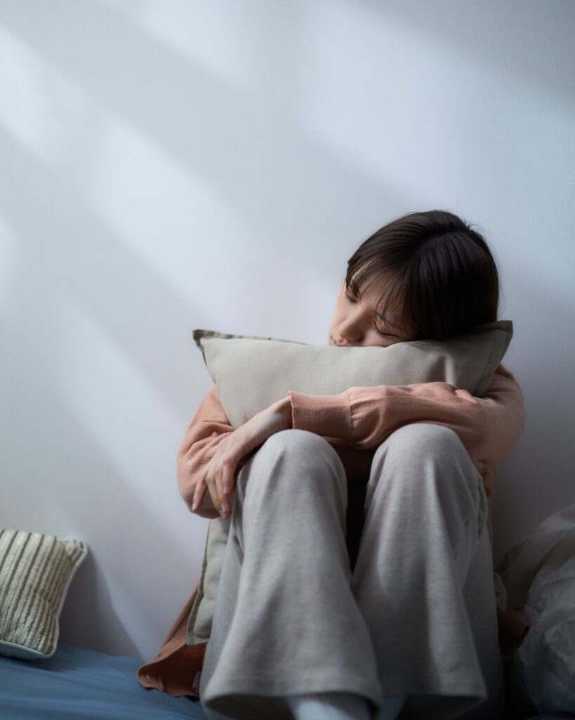 Midlife woman sitting in bed with eyes closed, head resting gently on a pillow; evoking rest, reflection, and hormonal recalibration.