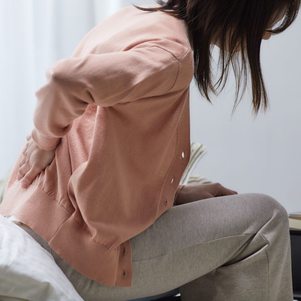 Midlife woman leaning forward slightly and holding her lower back, suggesting muscle pain or discomfort in the hips and lumbar region.