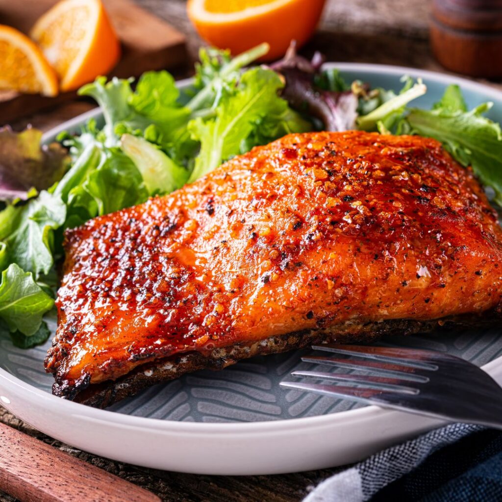 Orange salmon fillet with crispy skin, fresh greens, and citrus slices on a plate.