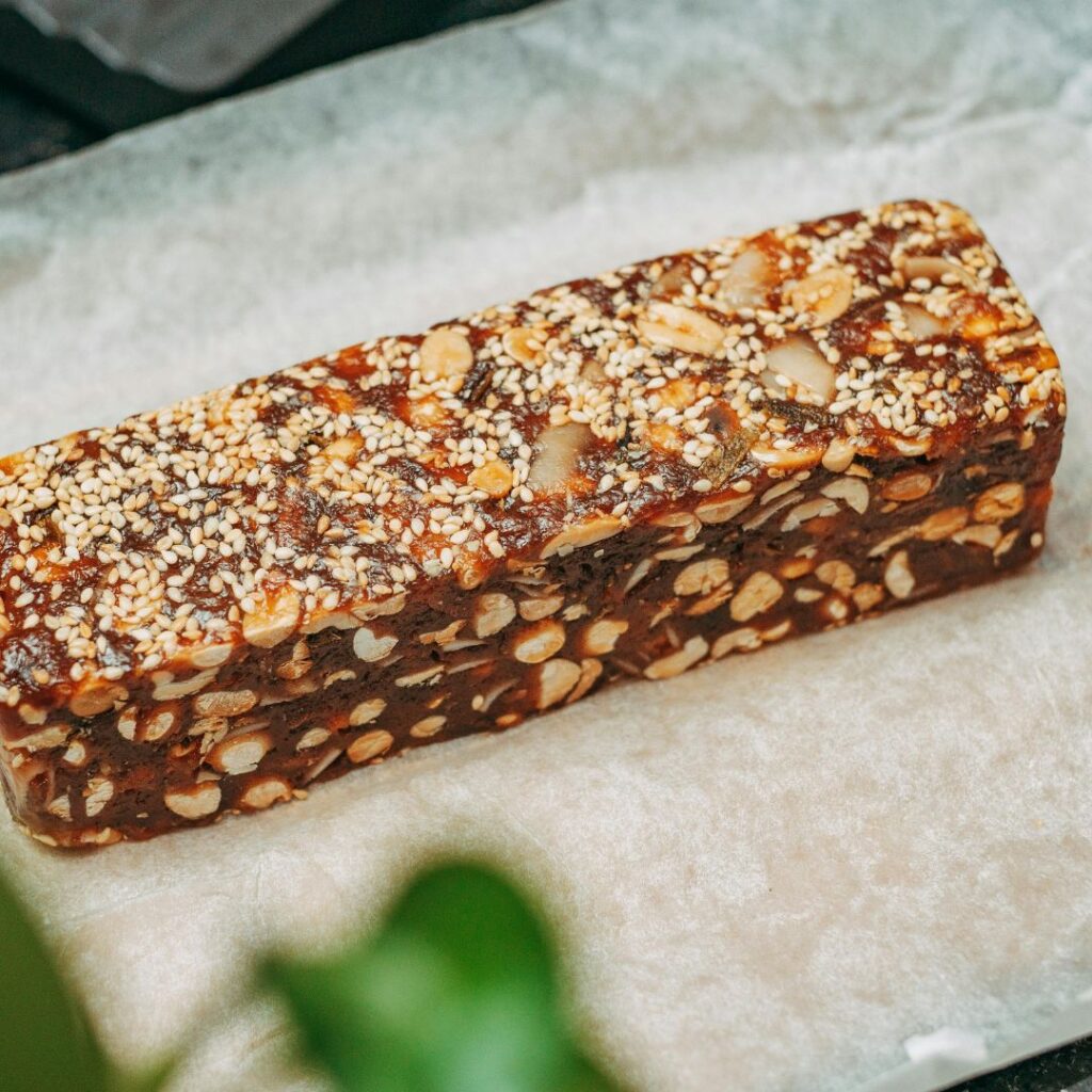 A rectangular protein bar showing visible sesame seeds on top and dense texture filled with chopped almonds and pumpkin seeds, ideal for midlife health.