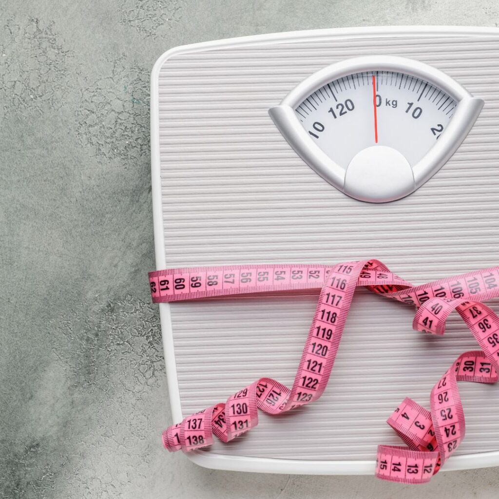 Close-up of a white analog scale and pink measuring tape on a gray surface, representing tracking weight during postmenopause.