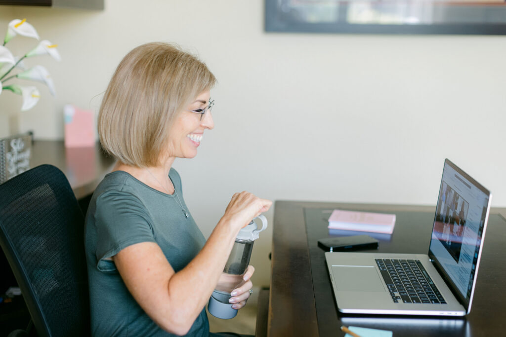 Holistic Nutritionist Lora Ulrich at her desk hosting an hosting an online consultation for midlife women with a water bottle in hand.