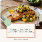 A grilled salmon fillet on a rustic plate, covered in mango-avocado salsa, sitting on a wooden table.