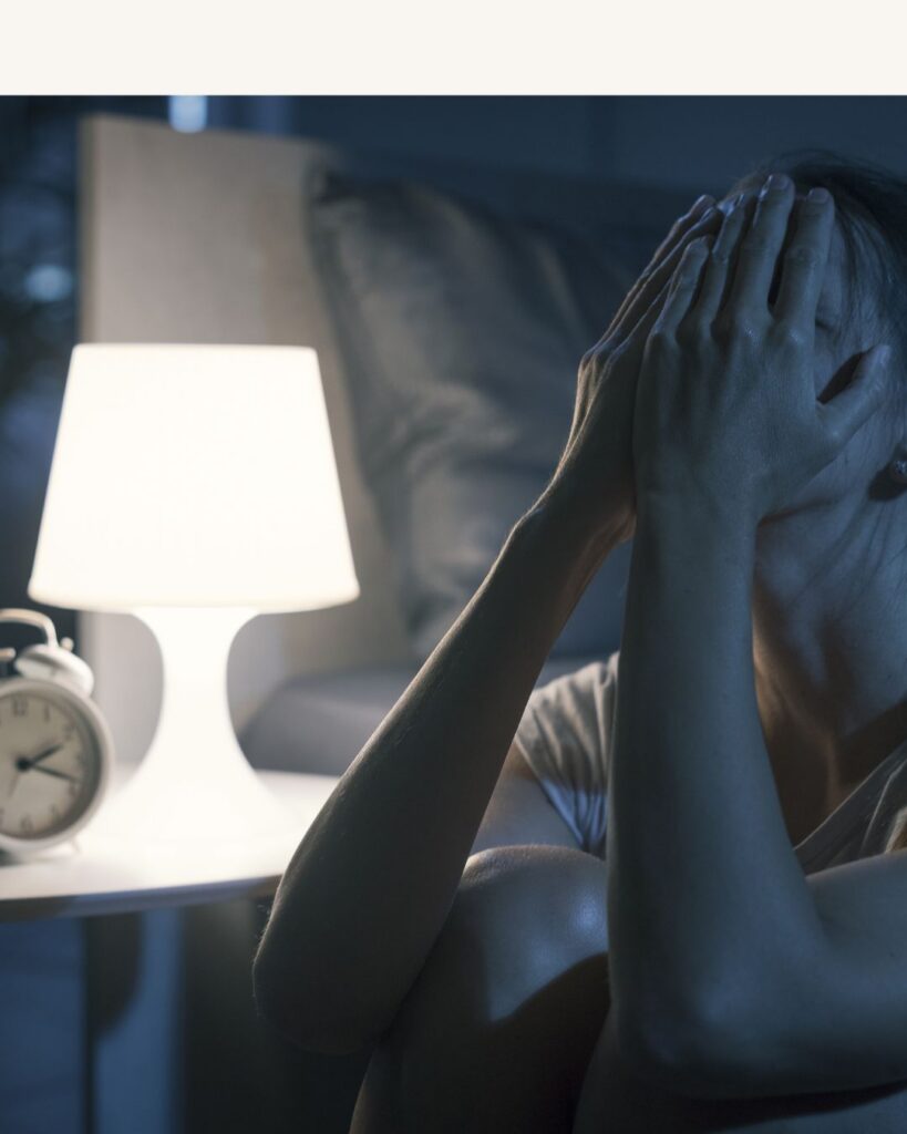 A tired woman sitting up in bed at night with her hands over her face, showing exhaustion from sleep loss during menopause.