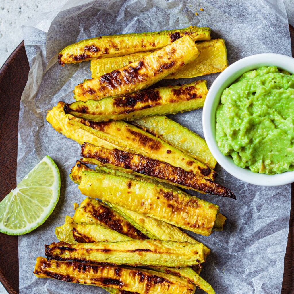 Golden crispy air fryer zucchini fries served on parchment paper with avocado dip and a lime wedge.