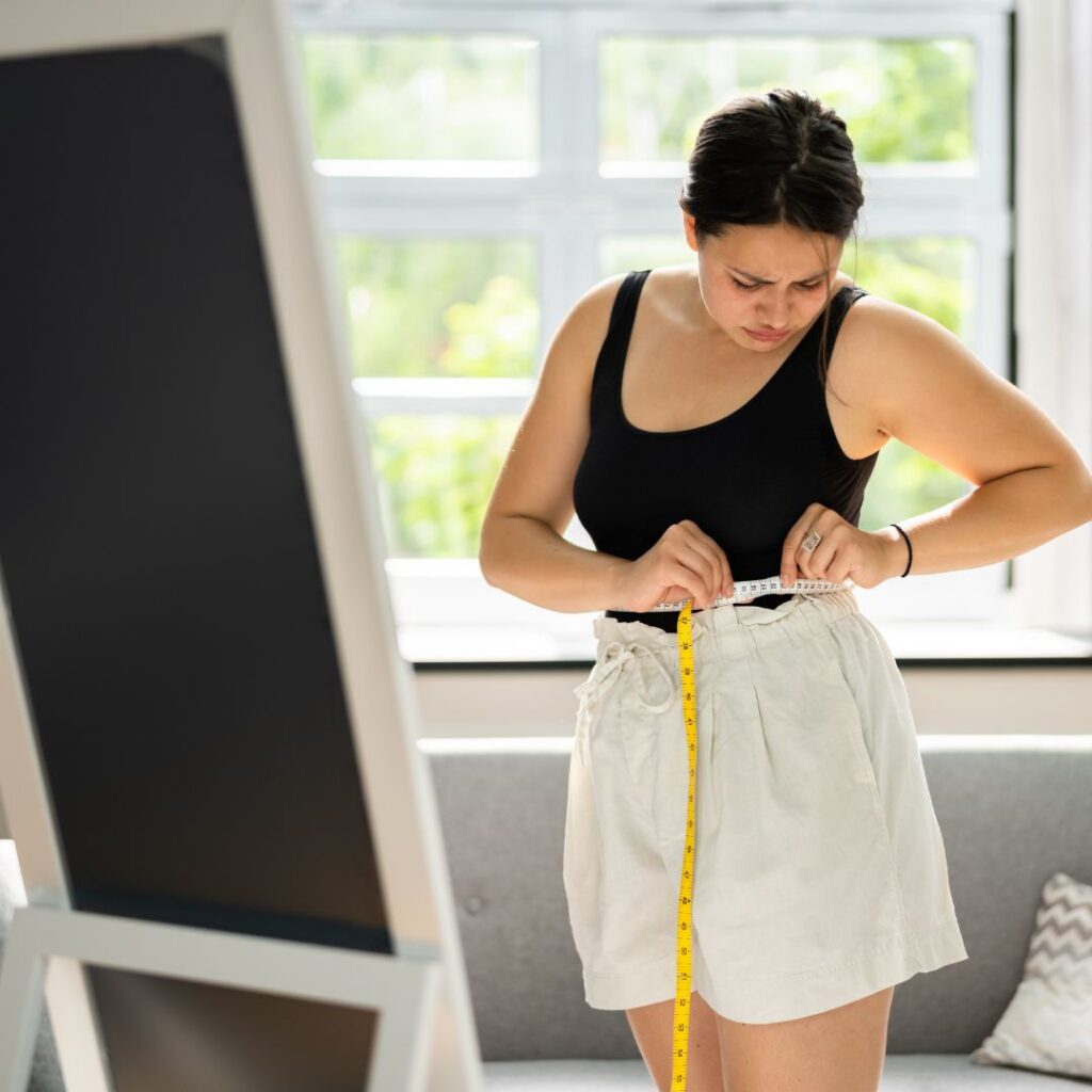 Frustrated woman measuring her waist with a tape measure in front of a mirror, illustrating perimenopause weight gain.