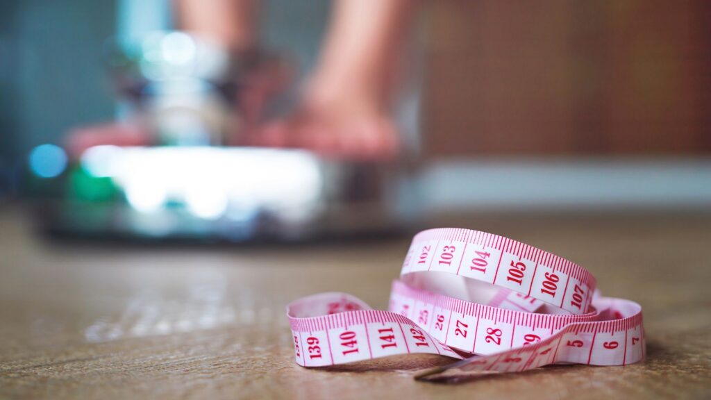 Pink measuring tape curled on the floor near a scale representing post-menopause weight gain and midsection body shifts.