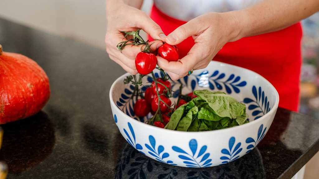 Hands preparing a healthy bowl with fresh cherry tomatoes and greens in a kitchen, representing blood sugar balancing meals for midlife health.
